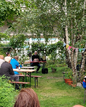 Im Hintergrund des Bildes ist Kolja Nixdorf zu sehen, der an einem Tisch sitzt und liest. Im Vordergrund sitzen Zuhörer und Zuhörerinnen an Biertischen und lauschen.