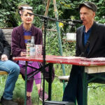 Der Kurator der "Epischen Nische", Claudius Hagemeister, sitzt mit der Autorin Oda Sajouz an einem Tisch im Garten der Kulturfabrik Moabit. Er spricht in ein Micro und eröffnet die Veranstaltung, indem er die Autorin und den Autoren Kolja Nixdorf kurz einführt.