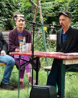 Der Kurator der "Epischen Nische", Claudius Hagemeister, sitzt mit der Autorin Oda Sajouz an einem Tisch im Garten der Kulturfabrik Moabit. Er spricht in ein Micro und eröffnet die Veranstaltung, indem er die Autorin und den Autoren Kolja Nixdorf kurz einführt.