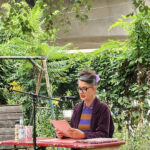 Oda Sajouz sitzt am Tisch im Garten der Kulturfabrik Moabit. In der Hand hält sie einige Seiten, von denen sie ihren Text abliest. Sie spricht in ein Micro.