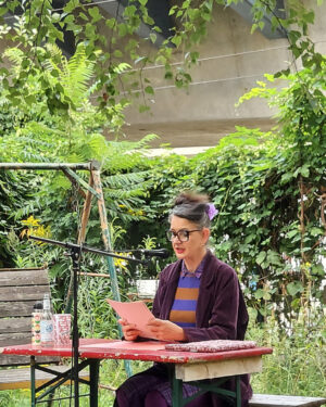Oda Sajouz sitzt am Tisch im Garten der Kulturfabrik Moabit. In der Hand hält sie einige Seiten, von denen sie ihren Text abliest. Sie spricht in ein Micro.