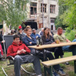 Das Publikum sitzt auf Bänken und Stühlen, im Hintergrund ist die Rückansicht der Kulturfabrik zu sehen.