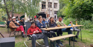 Das Publikum sitzt auf Bänken und Stühlen, im Hintergrund ist die Rückansicht der Kulturfabrik zu sehen.